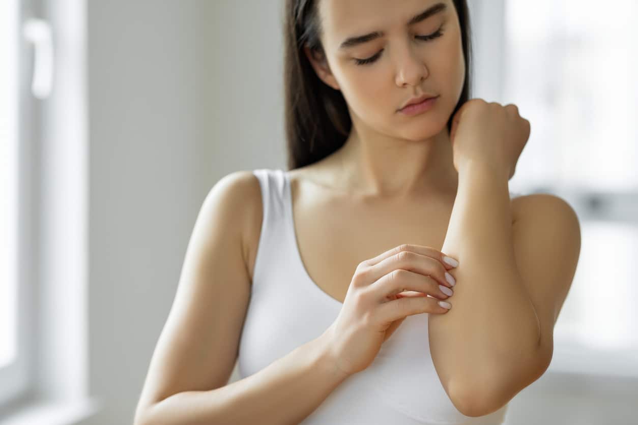 A Young Woman With Brown Hair In A White Tank Top Scratching Her Arm.